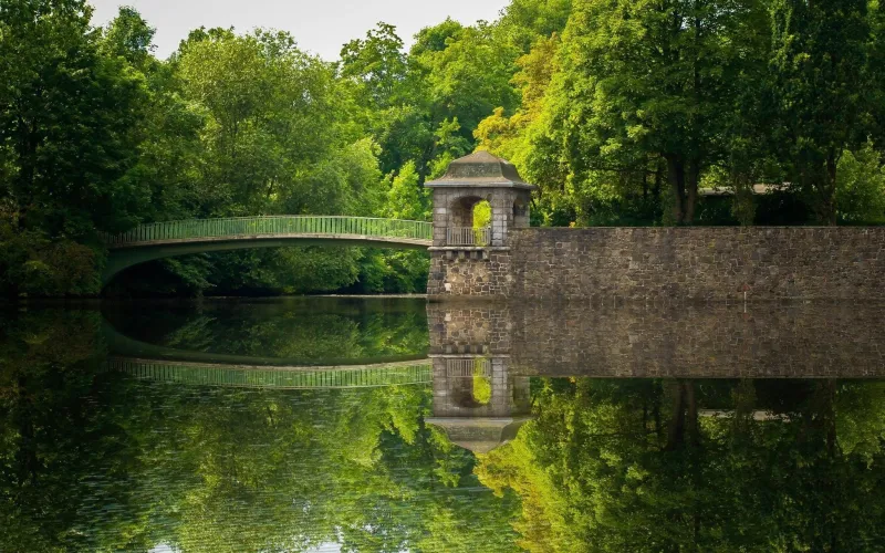 Teich mit Fußgängerbrücke, kleinem Steinturm und Bruchsteinmauer, umgeben von Laubbäumen, die sich im Wasser spiegeln